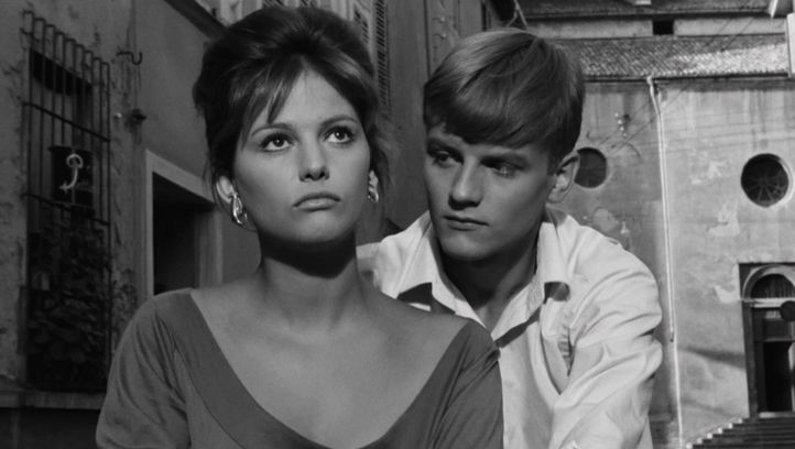 Film still from LA RAGAZZA CON LA VALIGIA: Claudia Cardinale, wearing a low-cut top, stands in the street and looks up at the sky. Behind her stands a teenager, looking at her.