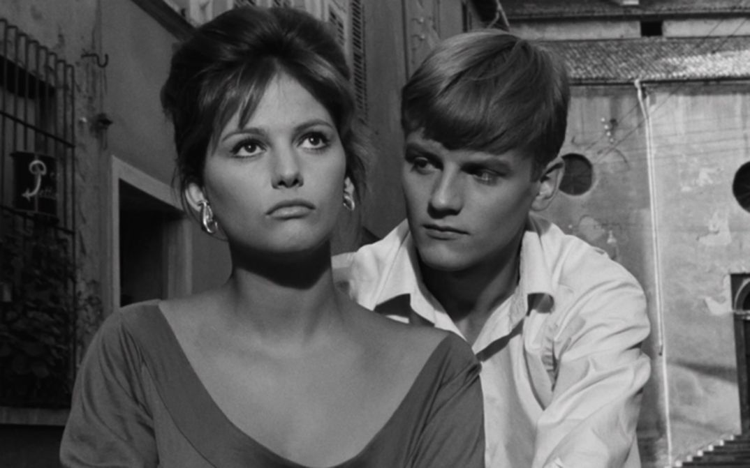 Film still from LA RAGAZZA CON LA VALIGIA: Claudia Cardinale, wearing a low-cut top, stands in the street and looks up at the sky. Behind her stands a teenager, looking at her.