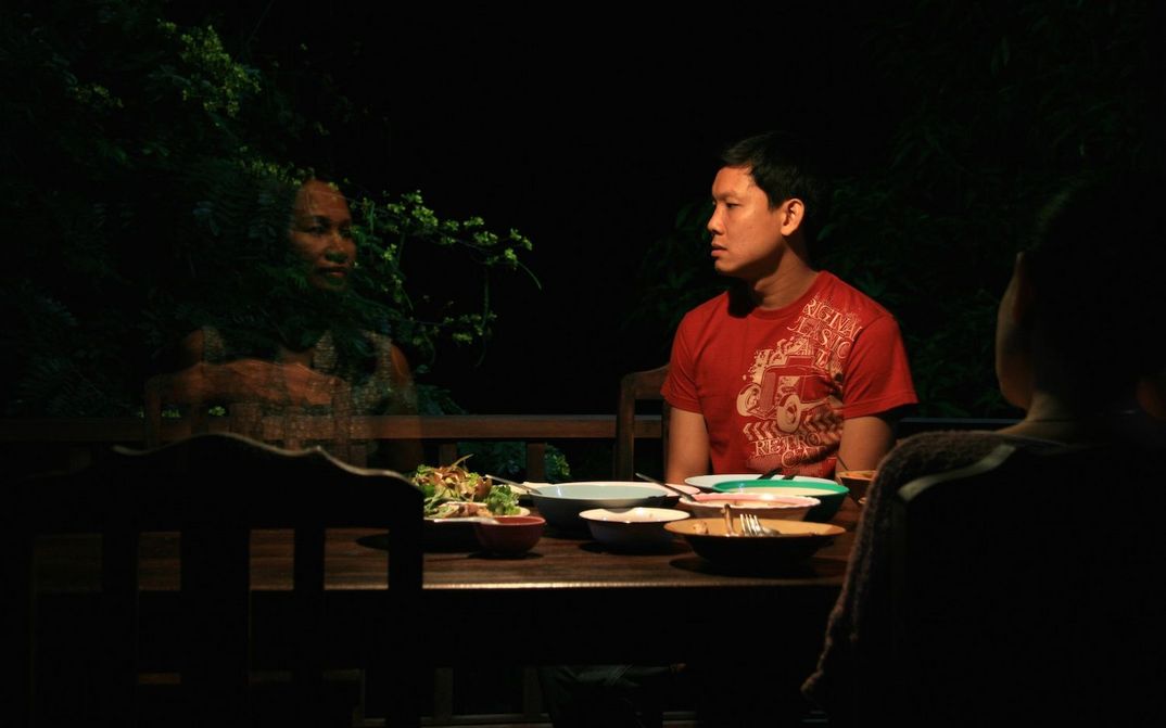 Film still from UNCLE BOONMEE WHO CAN RECALL HIS PAST LIVES: A man sits at a table in the darkness of the night and looks at a translucent woman.