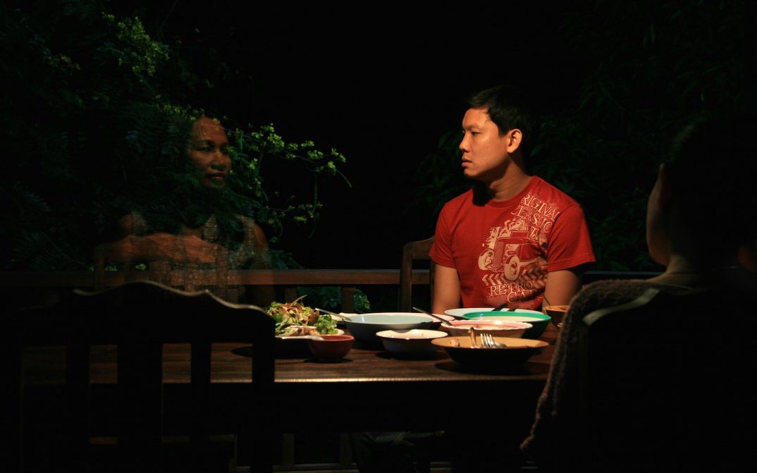 Filmstill aus UNCLE BOONMEE ERINNERT SICH AN SEINE FRÜHEREN LEBEN: Ein Mann sitzt in der nächtlichen Dunkelheit an einem Tisch und blickt auf eine durchscheinende Frau.
