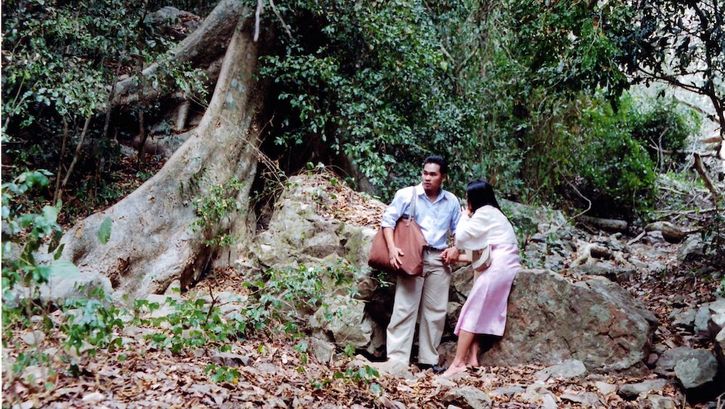 Film still from WORLDLY DESIRES: A man and a woman are standing in front of a large tree in a forest.