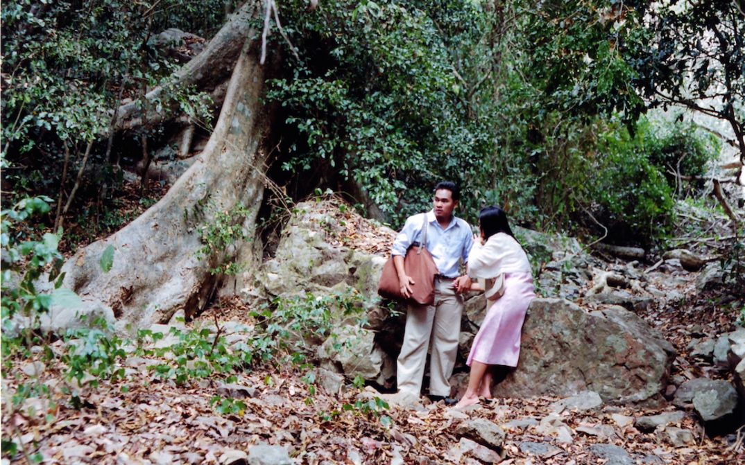 Filmstill aus WORLDLY DESIRES: Ein Mann und eine Frau stehen vor einem großen Baum in einem Wald.