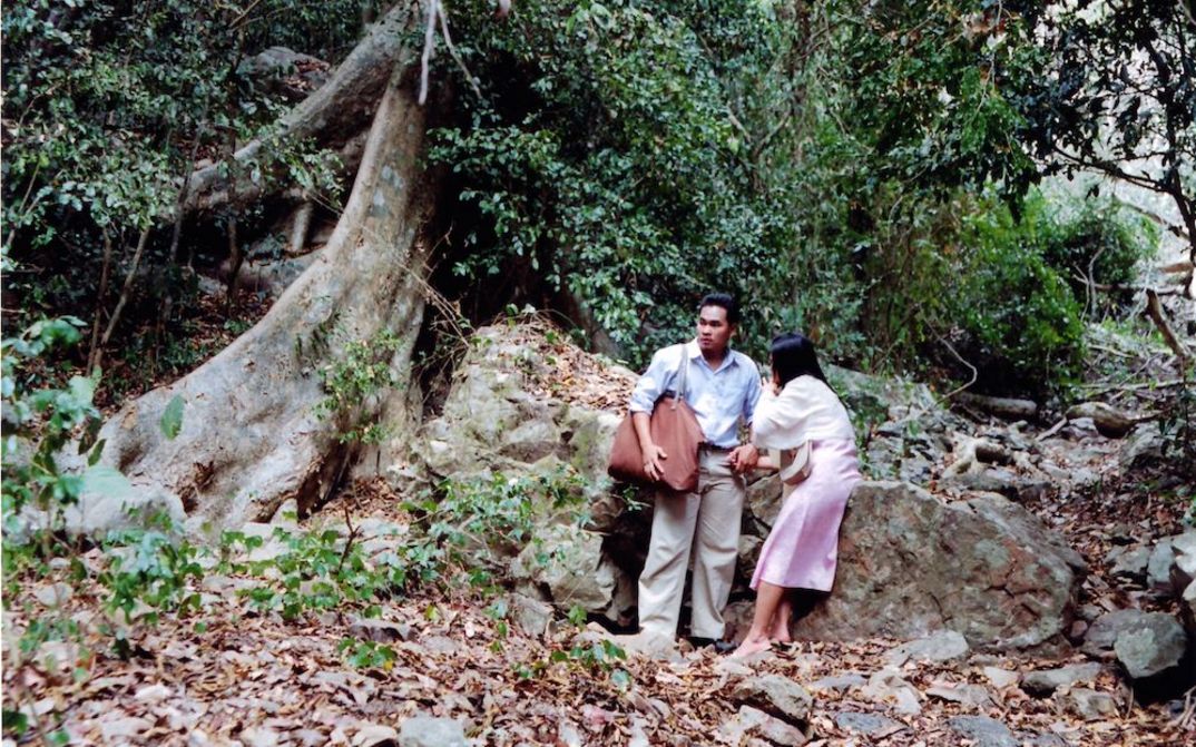 Film still from WORLDLY DESIRES: A man and a woman are standing in front of a large tree in a forest.