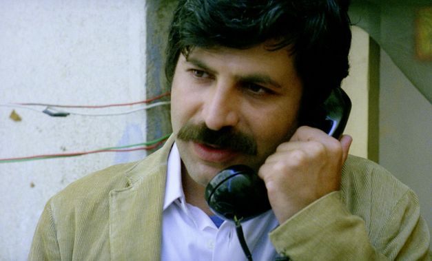 Still from the film "Beirut al lika (Beirut the Encounter)" by Borhane Alaouié. A man with a moustache in medium close-up speaks on a phone in front of a damaged exterior wall.
