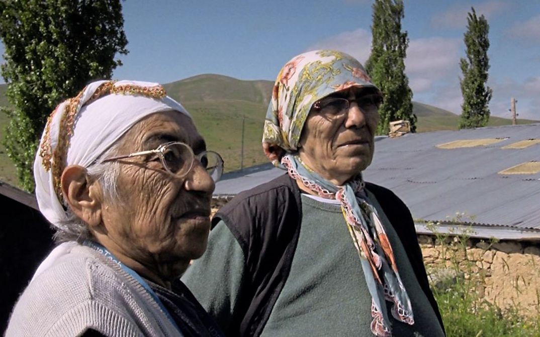 Filmstill aus „Dilim dönmüyor – Meine Zunge dreht sich nicht“ von Serpil Turhan. Zwei alte Frauen stehen auf einer Dorfstraße vor einem Lehmhaus.