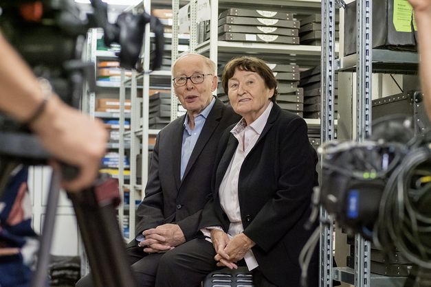 Still from the film "Come With Me to the Cinema – The Gregors" by Alice Agneskirchner. Two older people sitting in a film storeroom. At the left side of the frame we see a camera with the hand of the camera man.