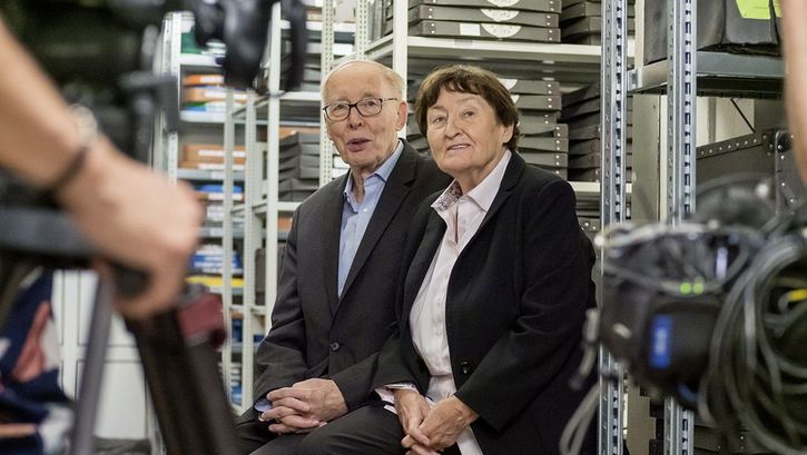 Still from the film "Come With Me to the Cinema – The Gregors" by Alice Agneskirchner. Two older people sitting in a film storeroom. At the left side of the frame we see a camera with the hand of the camera man.