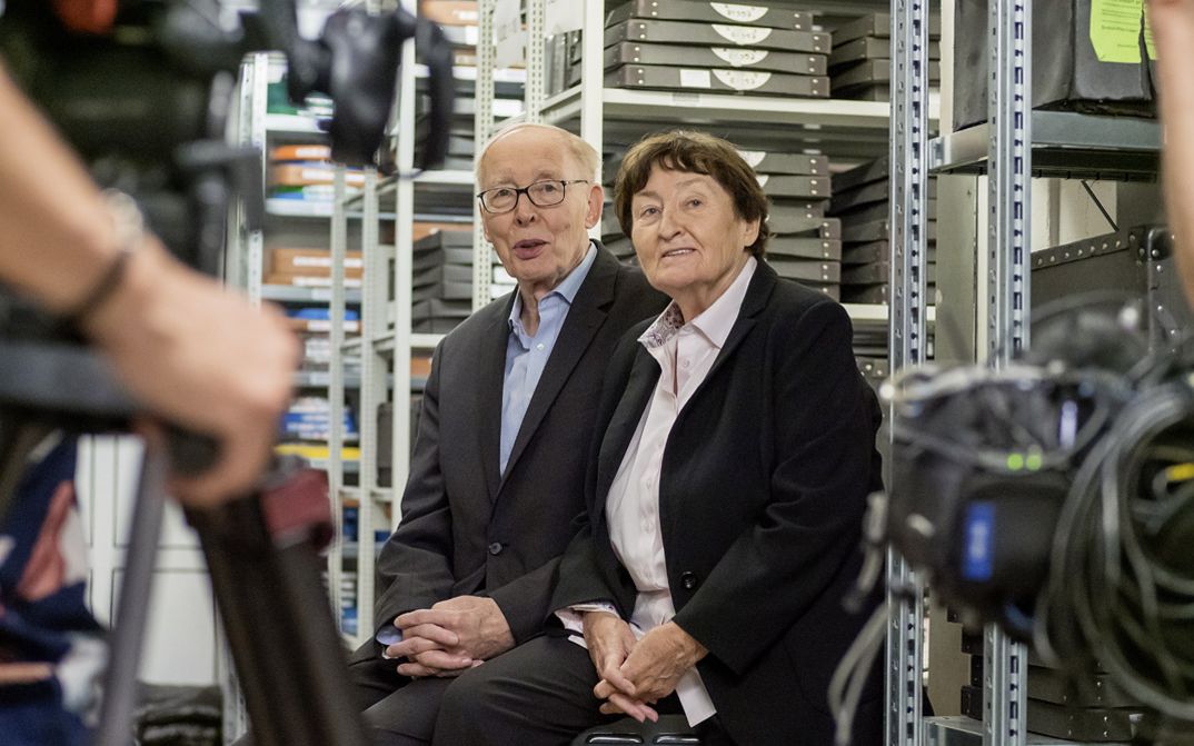Still from the film "Come With Me to the Cinema – The Gregors" by Alice Agneskirchner. Two older people sitting in a film storeroom. At the left side of the frame we see a camera with the hand of the camera man.