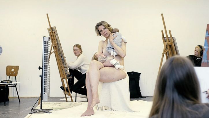 Filmstill aus "Reproduktion" von Katharina Pethke. Zu sehen ist eine nackte Frau mit zwei Babys im Arm, die auf einem Hocker sitzt. Um sie herum stehen Menschen an Staffeleien und zeichnen sie. 