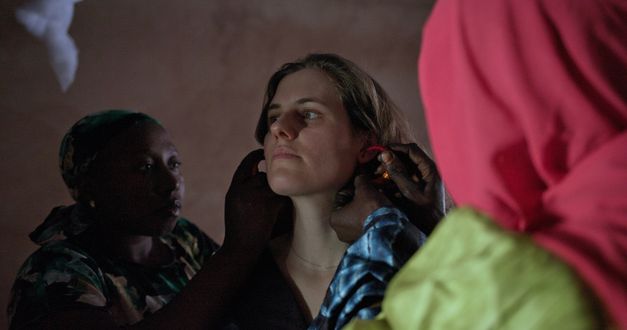 Film still from OUR TIME WILL COME by Ivette Löcker. It shows a woman being put on jewelry by two other women. 