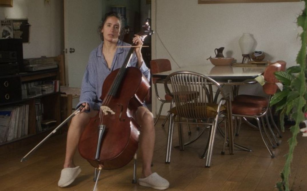 Person spielt Cello im Wohnzimmer, umgeben von Möbeln und Pflanzen.