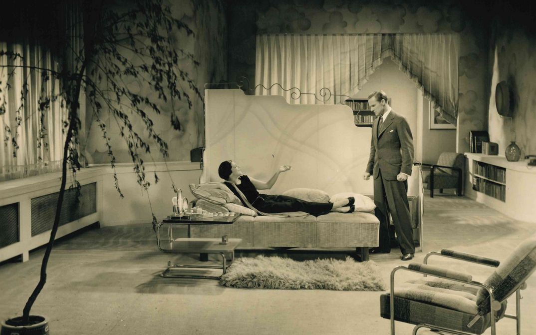 Filmstill aus STRAFSACHE VAN GELDERN: Ein Mann und eine Frau in einem glamourösen Salon. Sie legt auf einem Tagesbett und raucht eine Zigarette, er steht vor ihr.