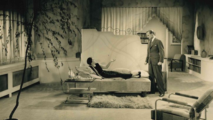 Filmstill aus STRAFSACHE VAN GELDERN: Ein Mann und eine Frau in einem glamourösen Salon. Sie legt auf einem Tagesbett und raucht eine Zigarette, er steht vor ihr.