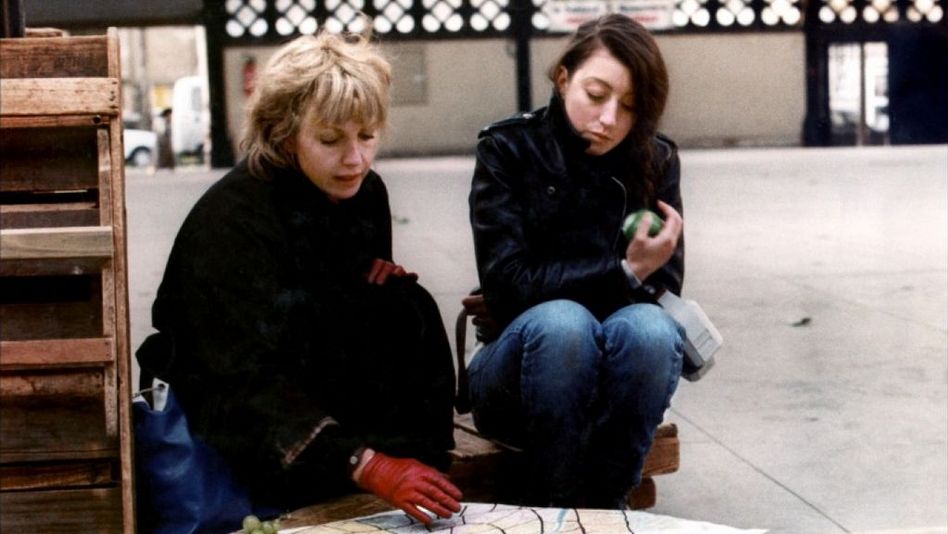 Film still from LE PONT DU NORD: Two women are sitting next to each other outdoors. They are looking at a cloth lying on a low table.
