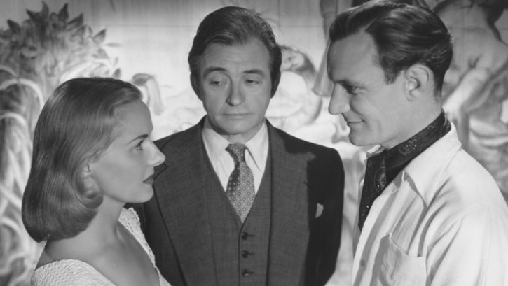 Filmstill aus THE PASSIONATE FRIENDS: Eine junge Frau und ein junger Mann blicken sich an. Sie sind im Profil zu sehen. Zwischen ihnen steht ein älterer Mann und blickt auf die Frau.