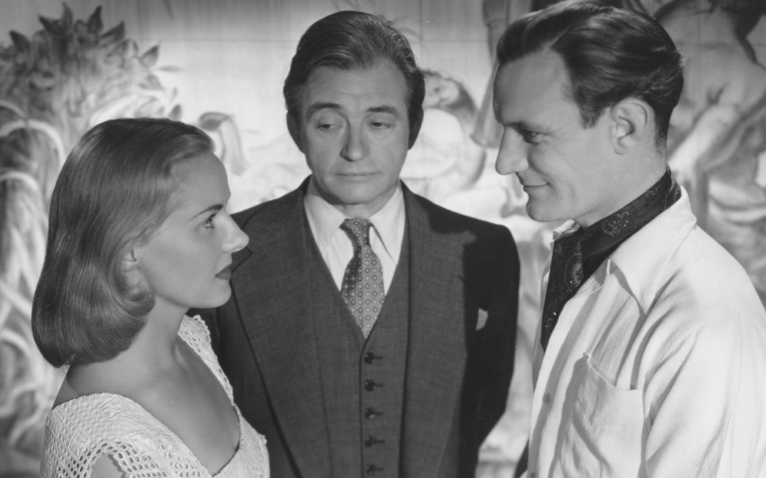 Filmstill aus THE PASSIONATE FRIENDS: Eine junge Frau und ein junger Mann blicken sich an. Sie sind im Profil zu sehen. Zwischen ihnen steht ein älterer Mann und blickt auf die Frau.