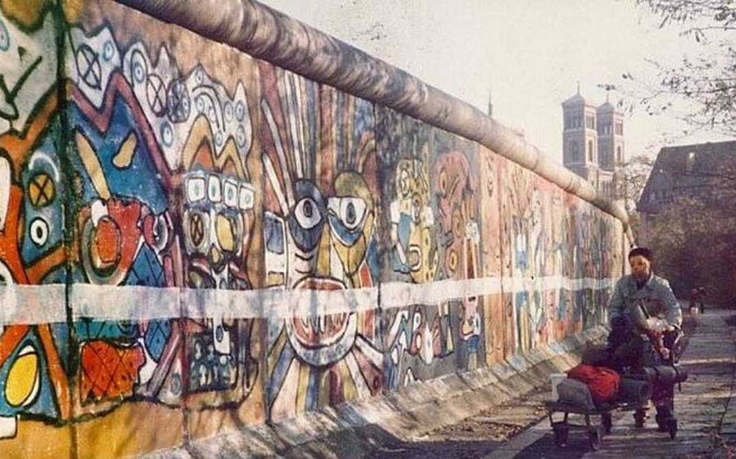 Filmstill aus STRICHE ZIEHEN: Zu sehen ist ein Stück der Berliner Mauer von westlicher Seite, sie ist bemalt und zusätzlich horizontal mit einer weißen Linie versehen.