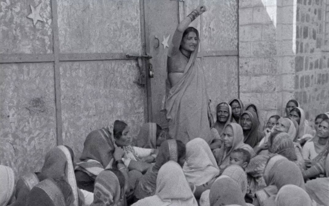 Filmstill aus dem Film „Tambaku Chaakila Oob Ali (Tobacco Embers)” vom Yugantar Film Collective. In Schwarz-Weiß sehen wir eine Frau vor einem steinernen Gebäude stehen und kämpferisch den Arm heben. Um sie herum gruppieren sich sitzende Frauen in Saris, unter ihnen auch ein Kind, und wenden sich ihr aufmerksam zu.