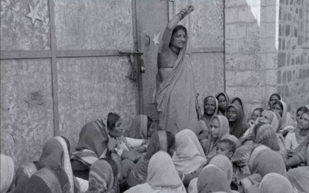 Filmstill aus dem Film „Tambaku Chaakila Oob Ali (Tobacco Embers)” vom Yugantar Film Collective. In Schwarz-Weiß sehen wir eine Frau vor einem steinernen Gebäude stehen und kämpferisch den Arm heben. Um sie herum gruppieren sich sitzende Frauen in Saris, unter ihnen auch ein Kind, und wenden sich ihr aufmerksam zu.
