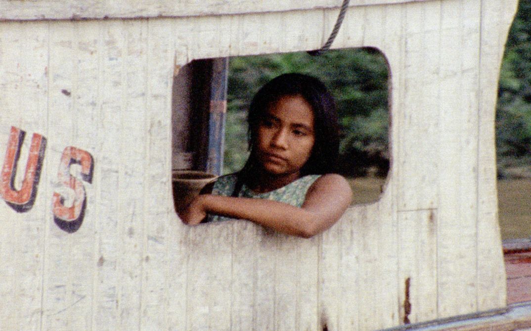 Film still from IRACEMA, UMA TRANSA AMAZÔNICA by Jorge Bodanzky and Orlando Senna. A young woman sits on the deck of a boat. Parts of an inscription, the letters US, can be seen on the outer paneling.