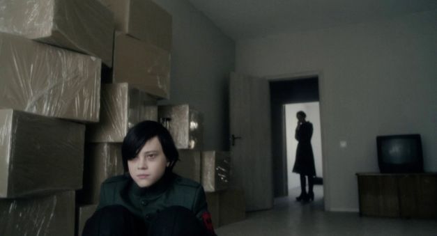 Filmstill aus CADET von Adilkhan Yerzhanov. Das Bild zeigt einen Jungen in Uniform, der auf dem Boden hockt, und die Silhouette einer Person im Hintergrund im Türrahmen.