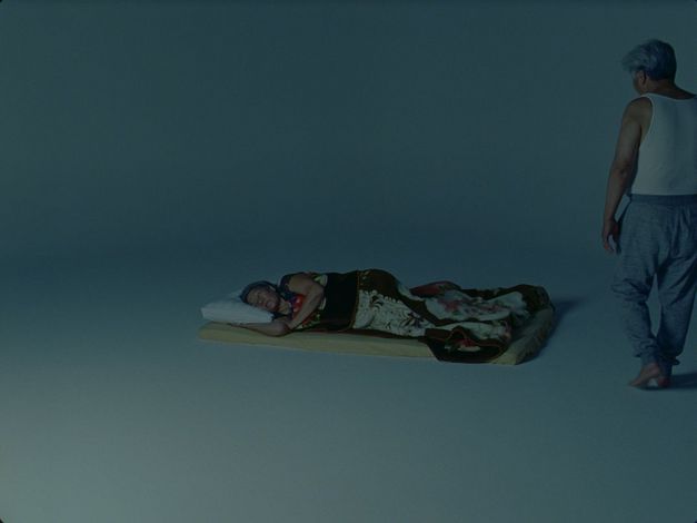 Filmstill aus dem Film „Dreams“ von Tenzin Phuntsog. Eine ältere Person schläft auf einer Matratze in einem sonst leeren Raum. Zu ihrer rechten steht eine grauhaarige Person in Unterhemd.
