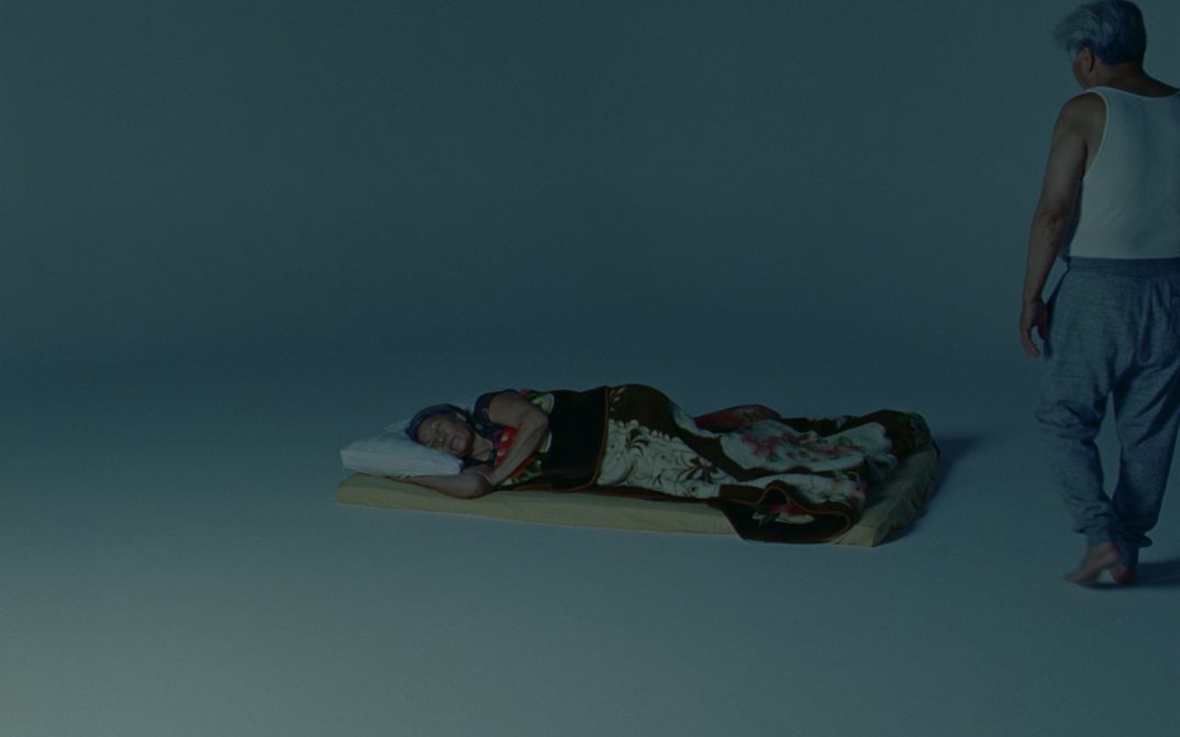 Film still from Tenzin Phuntsog’s „Dreams“. An elderly person sleeping on a matress in an otherwise empty room. To their right stands another person with grey hair, wearing a singlet.