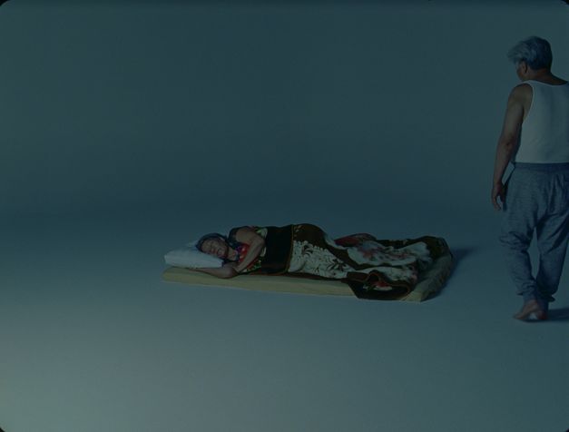 Film still from Tenzin Phuntsog’s „Dreams“. An elderly person sleeping on a matress in an otherwise empty room. To their right stands another person with grey hair, wearing a singlet.