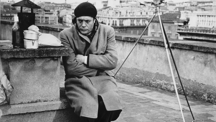 Film still from I SOLITI IGNOTI: Marcello Mastroianni sits on the roof of a building in a coat and cap.