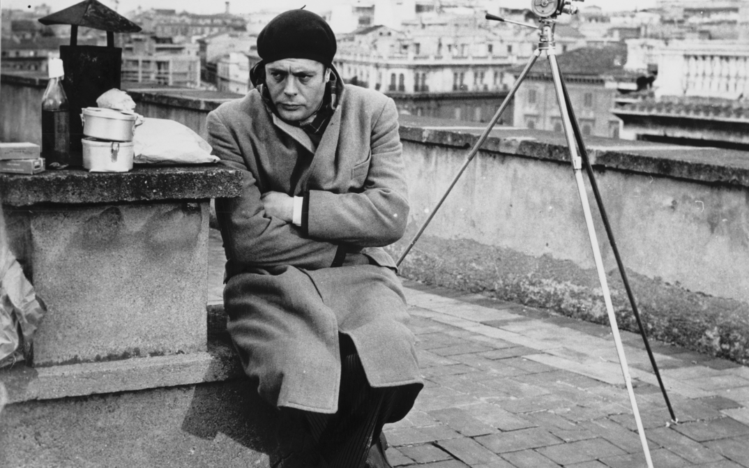 Film still from I SOLITI IGNOTI: Marcello Mastroianni sits on the roof of a building in a coat and cap.