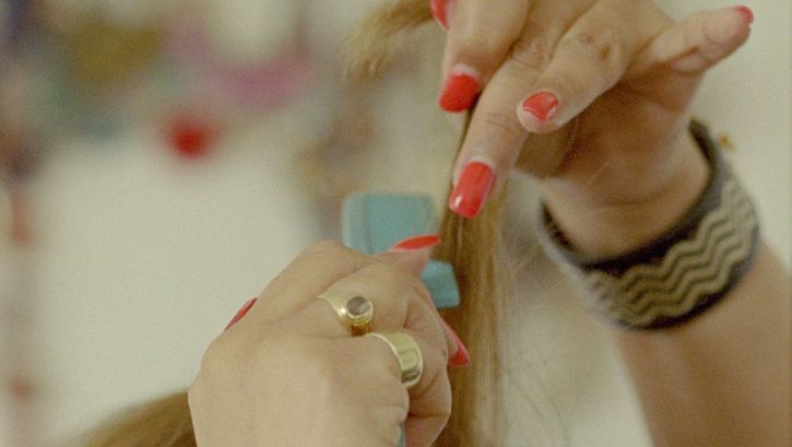 Still from the film "Mis Dos Voces" by Lina Rodriguez. We see a close-up of two hands with red painted fingernails running a blue comb through blonde hair. 