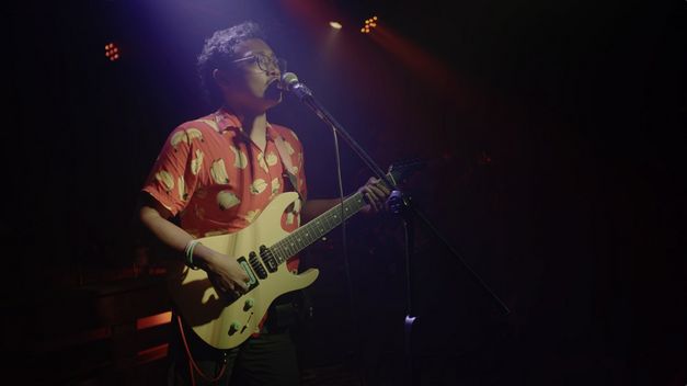 Film still from QUEER AS PUNK by Yihwen Chen. The image shows a person performing with a guitar and microphone in the spotlight.