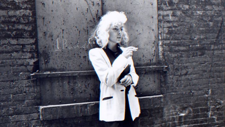 Filmstill from FOREIGNER:  Eine Faru mit blonden Haaren und weißem Jackett lehnt rauchend an einer Backsteinwand und blickt nachdenklich nach rechts.
