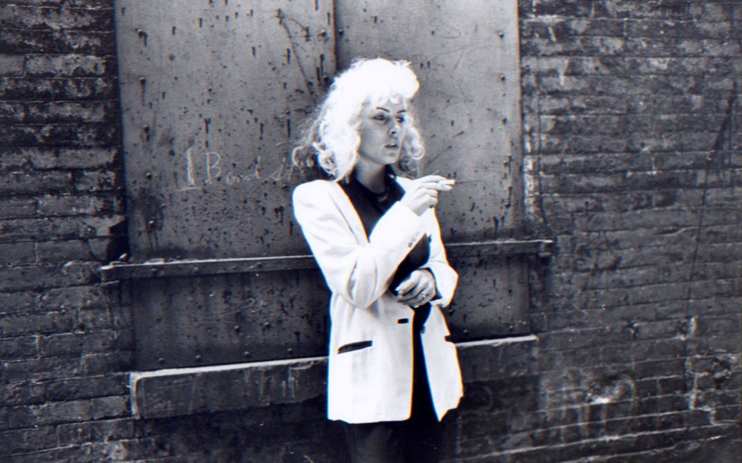 Filmstill from FOREIGNER:  Eine Faru mit blonden Haaren und weißem Jackett lehnt rauchend an einer Backsteinwand und blickt nachdenklich nach rechts.