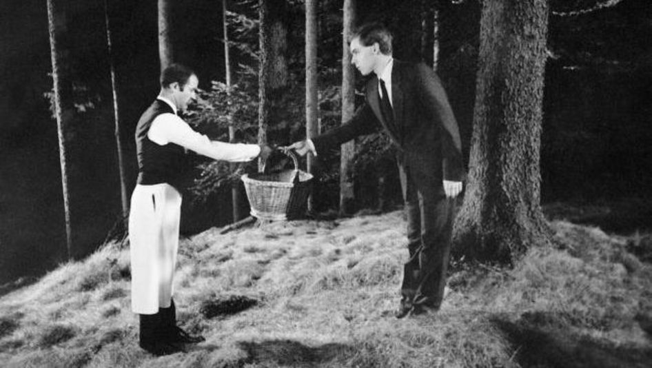Film still from KLASSENVERHÄLTNISSE: In the middle of a forest, a man wearing a waiter's apron hands a basket to a man in a suit.
