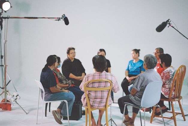 Menschen sitzen in einem Filmstudio im Kreis auf unterschiedlichen Stühlen. 