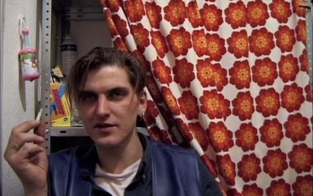 Film still from NICHTS FÜR DIE EWIGKEIT: A young man holding a cigarette looks into the camera. Behind him is a metal shelf, half-hidden by a floral curtain.