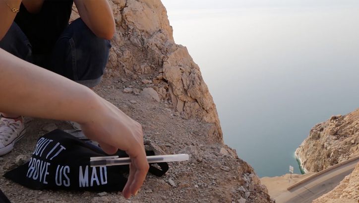 Filmstill aus dem Film "The Time That Separates Us" von Parastoo Anoushahpour. Arme und Unterkörper von zwei Personen, die auf einer Klippe hocken. Ein Stift in der Hand, eine Tasche auf dem Boden, unten eine Autostraße und blaues Meer.