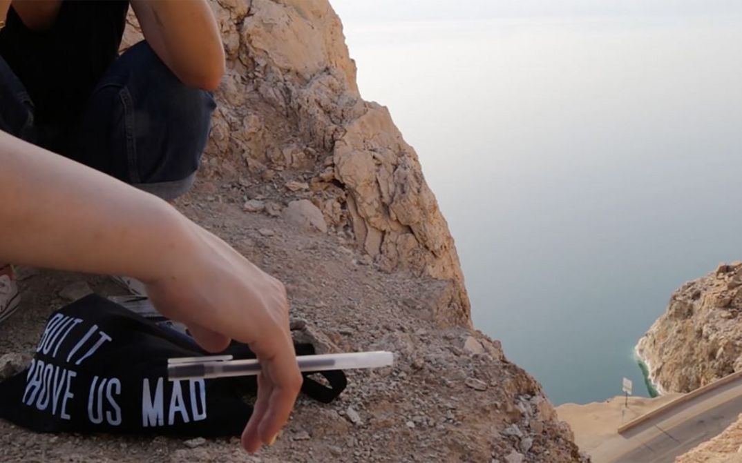 Filmstill aus dem Film "The Time That Separates Us" von Parastoo Anoushahpour. Arme und Unterkörper von zwei Personen, die auf einer Klippe hocken. Ein Stift in der Hand, eine Tasche auf dem Boden, unten eine Autostraße und blaues Meer.