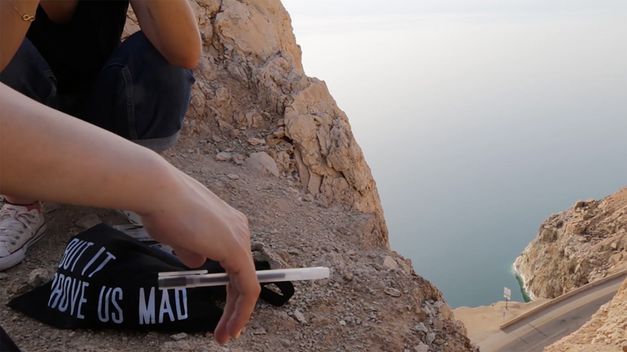 Film still from Parastoo Anoushahpour’s "The Time That Separates Us". Arms and lower body of two people sitting on a cliff. A pen in hand, a bag on the ground, below a highway and the blue sea.