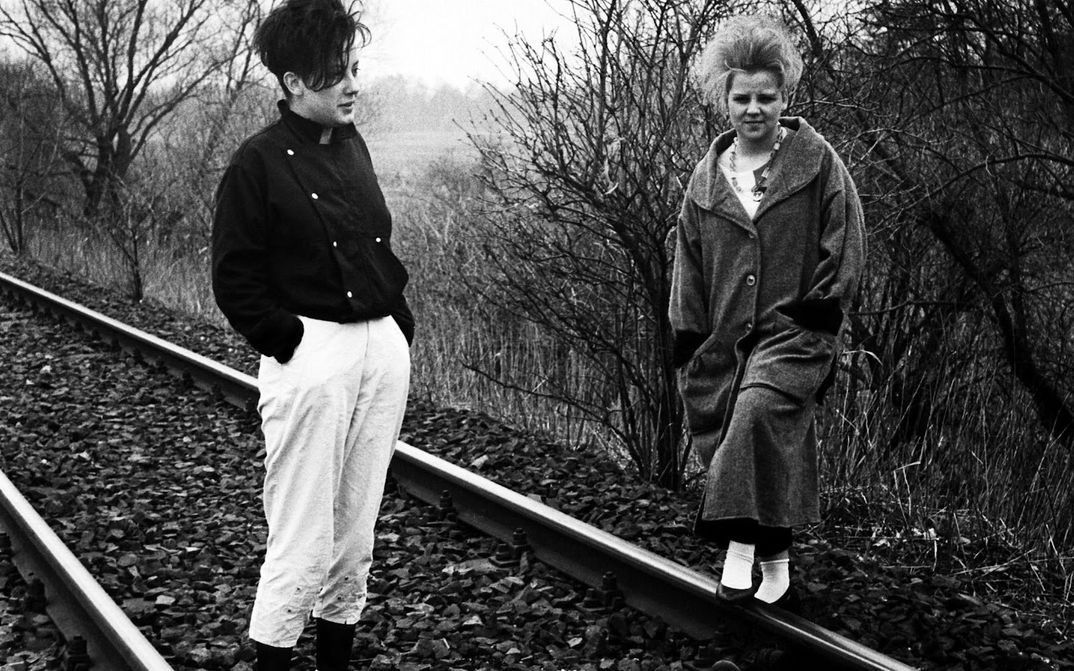 Film still from WINTER ADÉ: Two women stand on railway tracks. In the background is a field and trees. It looks dreary and grey.
