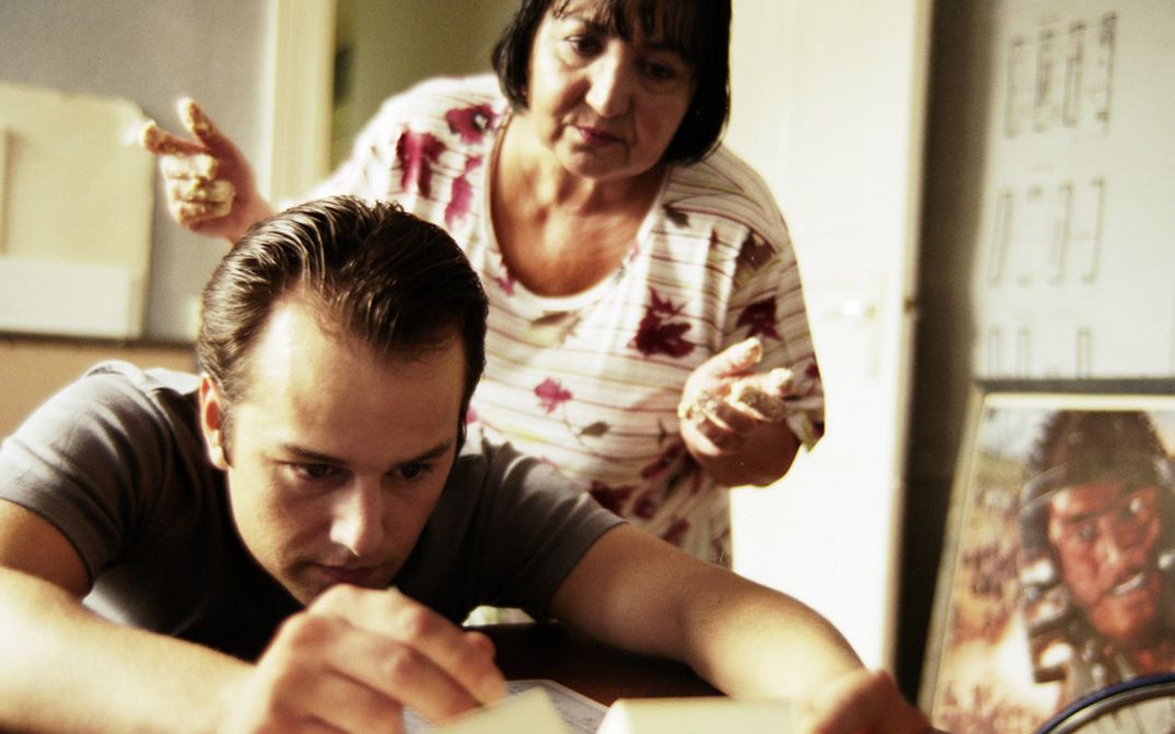  Filmstill aus "Fremd. Yaban." von Hakan Savaş Mican. Ein junger Mann beugt sich über einen Tisch, während ihm eine ältere Frau über die Schulter schaut. 