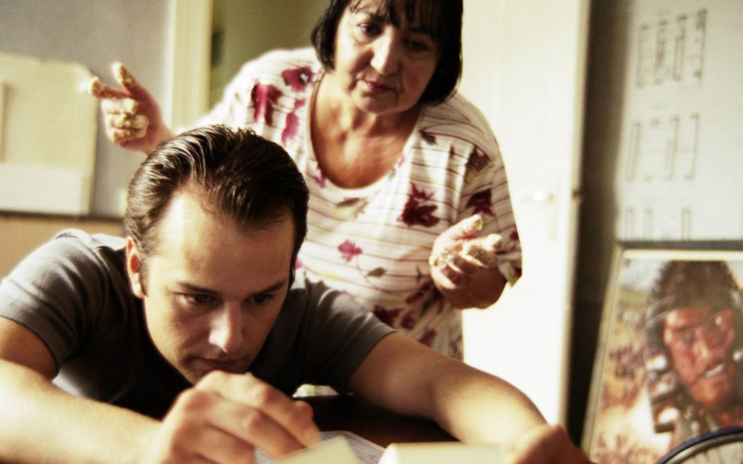  Filmstill aus "Fremd. Yaban." von Hakan Savaş Mican. Ein junger Mann beugt sich über einen Tisch, während ihm eine ältere Frau über die Schulter schaut. 