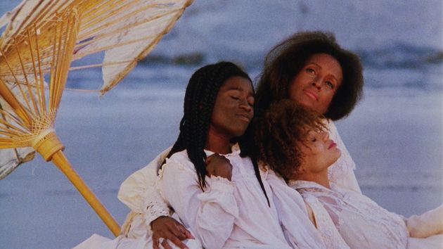 Still “Daughters of the Dust“. Three women wearing white are sitting on a beach, we can see the waves behind them. One woman is holding her arms around the other two. All three of them look dreamy. Behind them there is a parasol that is slightly broken.