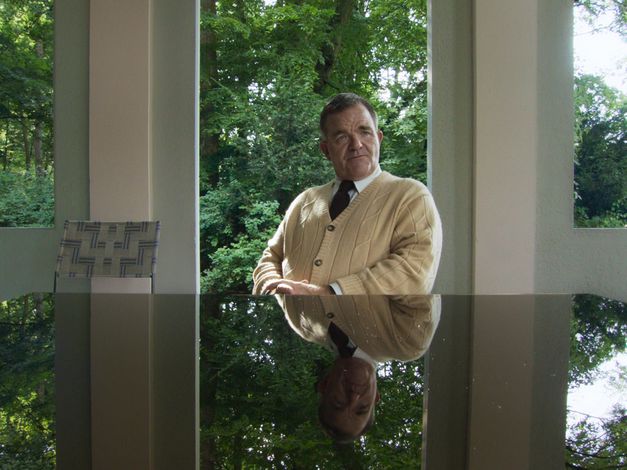 Still from „De Facto" von Selma Doborac. A man sitting at a black table, which mirrors his face and torso. Behind him you can see bushes and trees.