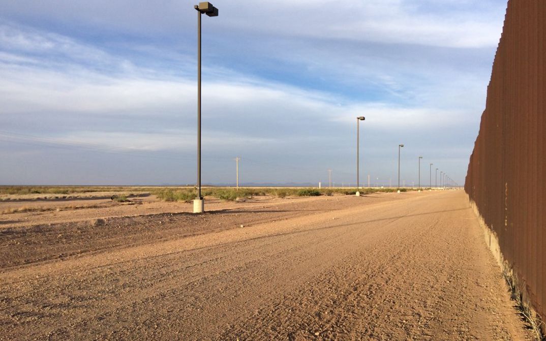 Film still from CROSSING COLUMBUS: A deserted, dusty road, the high border fence cut off to the left.