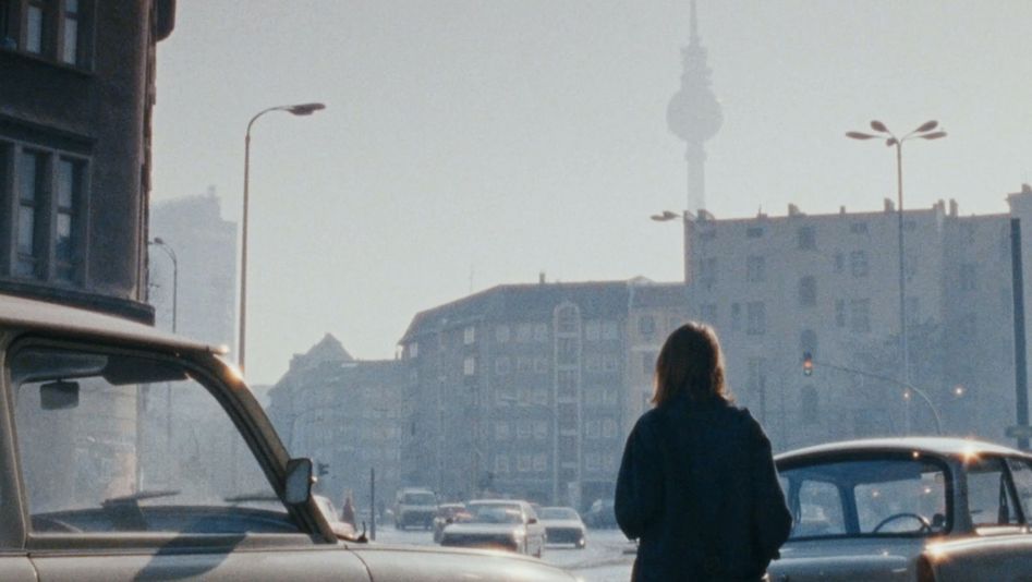Film still from OSTKREUZ: A young woman stands between cars on a street in Berlin. The television tower can be seen in the background.