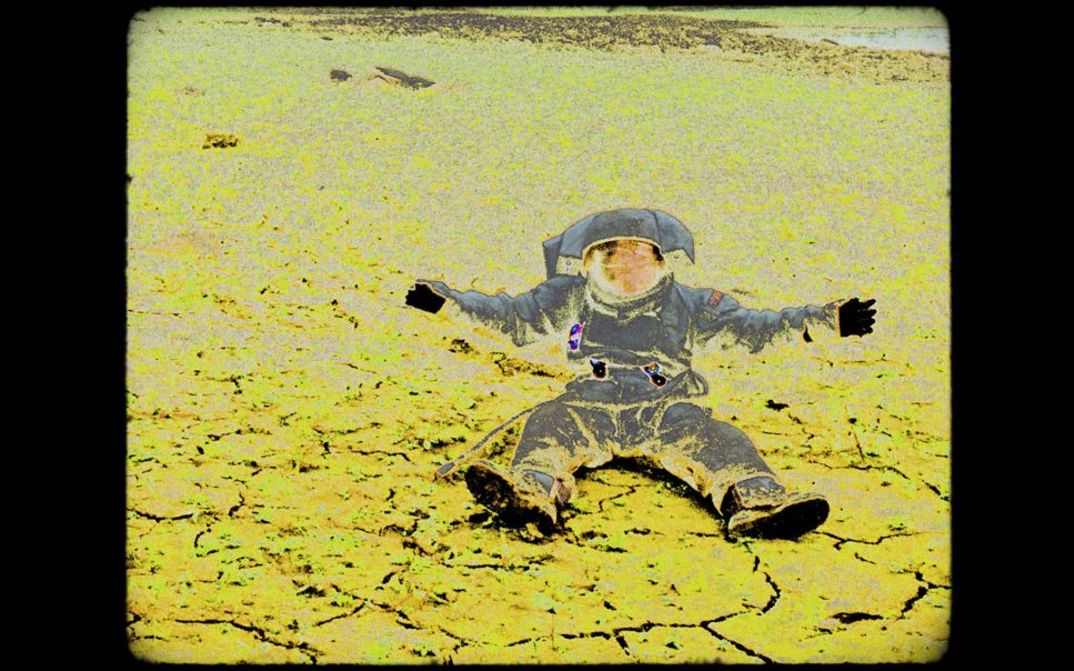 202301075_Poznámky_z_Eremocénu_1 An Astronauten in a space suit sitting with open arms in a deserted landscaped; the image looks like a negative print with apparent image interferences.