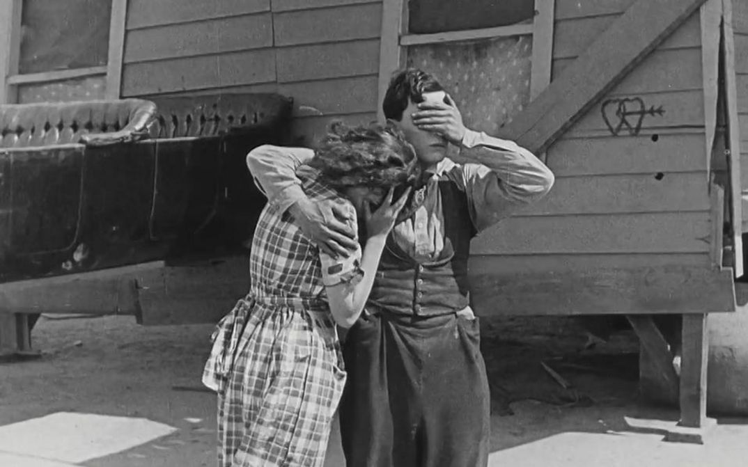 Filmstill aus ONE WEEK: Ein Mann und eine Frau stehen vor einem einfachen Holzhaus. Er hat den Arm um sie gelegt, beide halten ihre Hände vor die Augen.