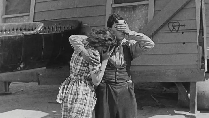 Film still from ONE WEEK: A man and a woman are standing in front of a simple wooden house. He has his arm around her, both are holding their hands in front of their eyes.